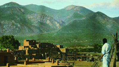 A person stands on a rooftop overlooking adobe buildings and wooden structures, with forested mountains in the background under a clear sky. [gen-ai]