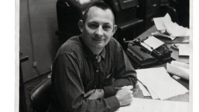 A man sits at a cluttered desk with papers, a typewriter, and office machines in the background, looking at the camera and smiling. [gen-ai]