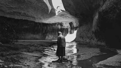 A person stands in a rocky canyon with high, curving walls and a narrow stream, illuminated by sunlight streaming through an opening above. [gen-ai]