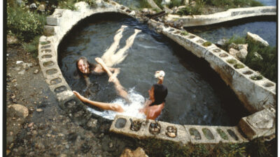 Two people relax in a small, concrete hot spring pool surrounded by rocks and greenery. [gen-ai]