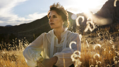 A woman in a white blouse sits in a sunlit field with tall grasses, mountains in the background, and the sun shining behind her. [gen-ai]
