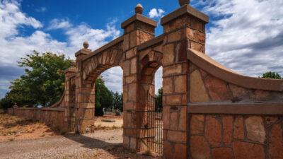 A stone archway entrance with a metal gate set in a brick and stone wall, surrounded by trees and under a partly cloudy sky. [gen-ai]