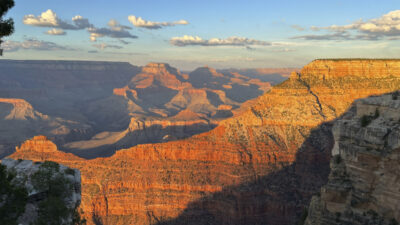 The Grand Canyon at sunset, with layered red-orange rock formations and deep shadows under a clear blue sky with scattered clouds. [gen-ai]
