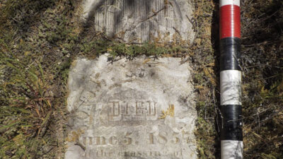 A weathered gravestone lies partially covered by grass, with two black, white, and red measuring rods placed alongside it for scale. [gen-ai]