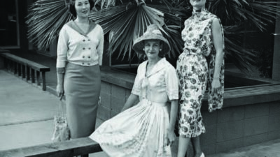 Three women in vintage clothing pose outdoors; one stands in a pencil skirt, one kneels in a wide-brimmed hat and dress, and the third stands in a floral dress and heels near palm leaves. [gen-ai]