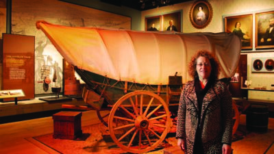 A woman stands in front of a covered wagon exhibit in a museum with framed portraits and historical displays in the background. [gen-ai]