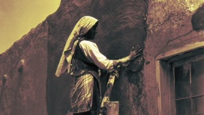 Two people apply adobe plaster to a wall; one stands on a ladder, while the other works on the roof. A window is visible on the right side of the building. [gen-ai]