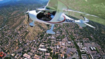 A small white airplane flies over a city, with buildings, roads, and greenery visible below. The pilot is seen through the cockpit window. [gen-ai]