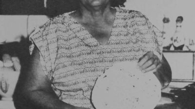 A woman standing in a kitchen holds a round, flat food item, possibly a tortilla, in front of her chest. [gen-ai]