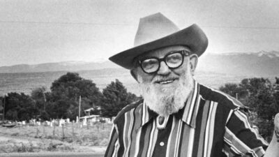 An older man with a white beard, glasses, and a cowboy hat stands outdoors in front of a rural landscape, smiling and holding a camera. [gen-ai]