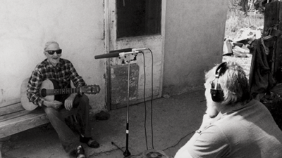 An older man with a guitar sits on a bench outside as another person with headphones records him using a reel-to-reel recorder and a microphone. [gen-ai]