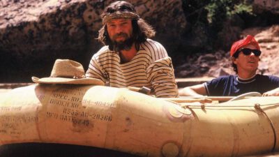 Two people ride an inflatable raft on a river; one wears a striped shirt and headband, the other a red bandana and sunglasses. A hat rests on the raft’s edge. Rocky terrain is visible in the background. [gen-ai]