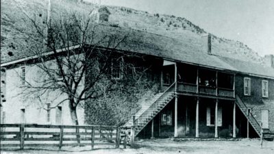 Black-and-white photo of a two-story adobe building with exterior stairs, a porch, and a fence in front; barren tree and hills in the background. [gen-ai]