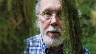 An older man with glasses and a white beard, wearing a blue plaid shirt, stands outdoors among green moss-covered branches. [gen-ai]