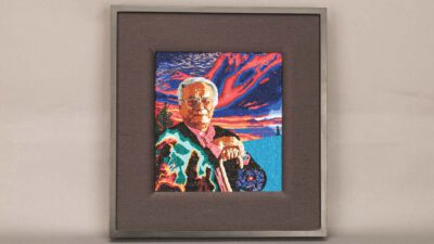 A framed needlepoint artwork depicts an elderly person in colorful clothing holding a round object, set against a vibrant sunset sky and landscape background. [gen-ai]
