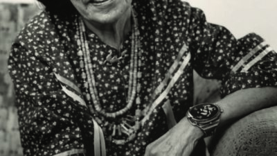An older woman with glasses and dark hair sits on a patterned chair, wearing a patterned blouse, necklaces, bracelets, and rings, looking at the camera with a gentle expression. [gen-ai]
