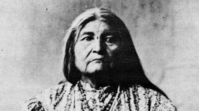 Black and white portrait of an older Indigenous woman with long hair, wearing patterned clothing and beaded necklaces, looking directly at the camera. [gen-ai]