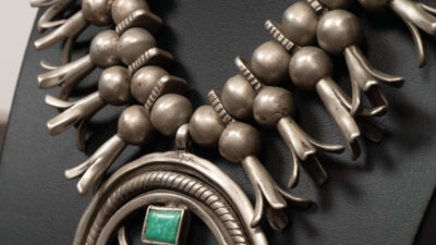 A silver necklace with large round beads and squash blossom motifs, featuring a central circular pendant with two turquoise stones, displayed on a black stand. [gen-ai]