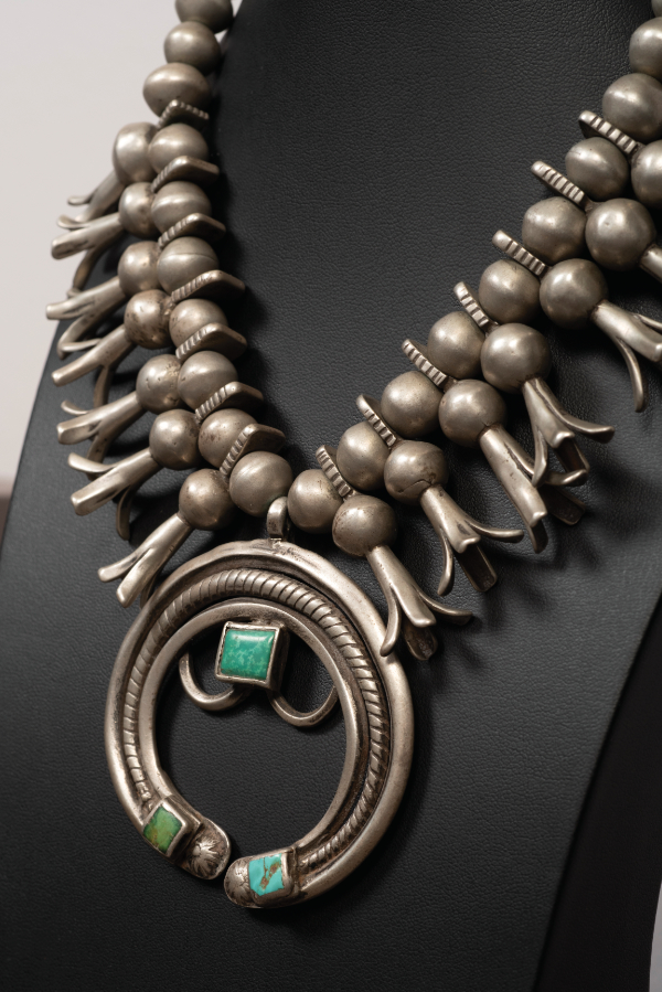 A silver necklace with large round beads and squash blossom motifs, featuring a central circular pendant with two turquoise stones, displayed on a black stand. [gen-ai]