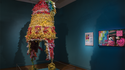 A large, colorful piñata sculpture displayed in a museum gallery with blue walls, next to an information plaque and a digital screen showing exhibit details. [gen-ai]