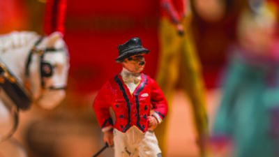 A close-up of a painted figurine in a red jacket, black boots, and hat holding a stick, with other colorful figurines and a white horse in the blurred background. [gen-ai]