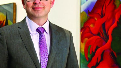 A man in a gray suit and purple tie stands in an art gallery next to a colorful abstract painting with red, green, and blue shapes. [gen-ai]