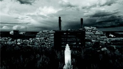 A stone grave marker stands in front of a crumbling stone wall, with a cloudy sky and rural landscape in the background. [gen-ai]
