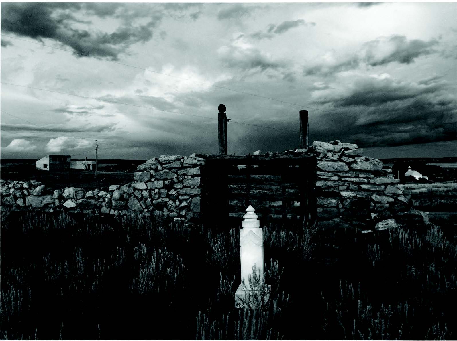 A stone grave marker stands in front of a crumbling stone wall, with a cloudy sky and rural landscape in the background. [gen-ai]