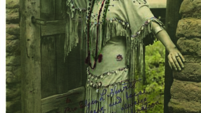 A woman in traditional Native American attire stands at the doorway of an adobe building, wearing a fringed dress, beaded accessories, and moccasins. [gen-ai]