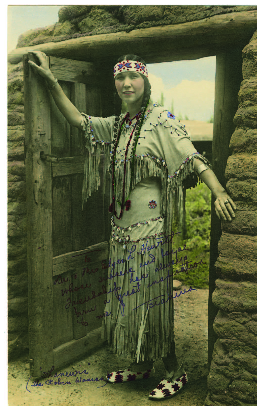 A woman in traditional Native American attire stands at the doorway of an adobe building, wearing a fringed dress, beaded accessories, and moccasins. [gen-ai]