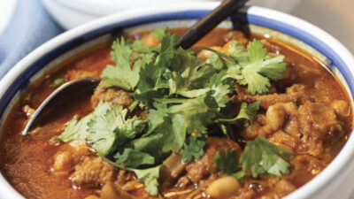 A bowl of red stew with chunks of meat and hominy, topped with fresh cilantro, with a spoon resting inside. [gen-ai]