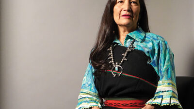 Woman with long dark hair sits against a plain background wearing a turquoise patterned blouse, black dress with red and green trim, and silver jewelry. [gen-ai]
