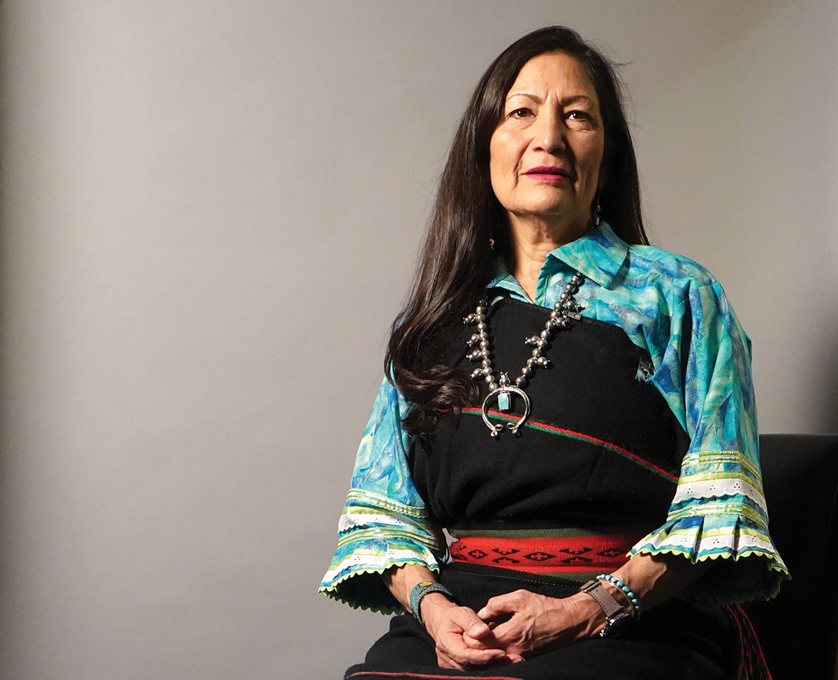Woman with long dark hair sits against a plain background wearing a turquoise patterned blouse, black dress with red and green trim, and silver jewelry. [gen-ai]