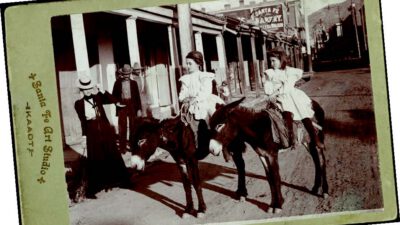 Two young girls in dresses sit on donkeys in a dirt street, with two adults standing nearby and buildings visible in the background. [gen-ai]