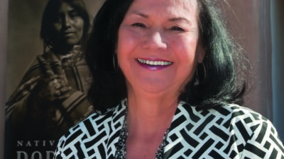 A woman with dark hair smiles outdoors in front of a poster featuring a person in traditional attire. She wears a patterned jacket and hoop earrings. [gen-ai]