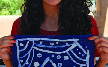 A smiling person with curly hair holds up a blue and white artwork depicting a house with a snowy scene. [gen-ai]