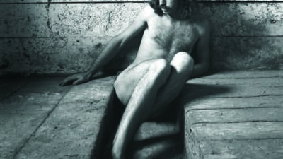 A nude person with long hair sits on a worn wooden floor, leaning against a textured wall in a dimly lit room. [gen-ai]