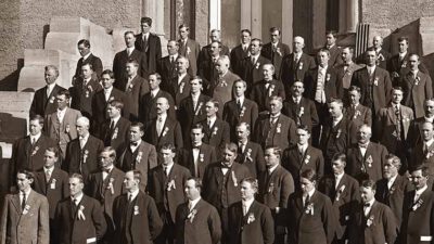 A large group of men in suits with ribbons on their lapels stand on stone steps outside a building, posing for a formal group photograph. [gen-ai]