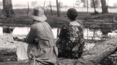 Two women sit on a fallen tree beside a pond, facing away from the camera in an outdoor, rural setting. [gen-ai]