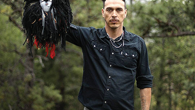 A man stands outdoors holding up a handmade mask adorned with feathers and face paint, surrounded by trees. [gen-ai]