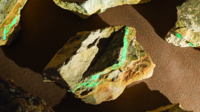 Several rough, jagged rocks with green mineral streaks rest on a brown textured surface, casting distinct shadows under angled lighting. [gen-ai]