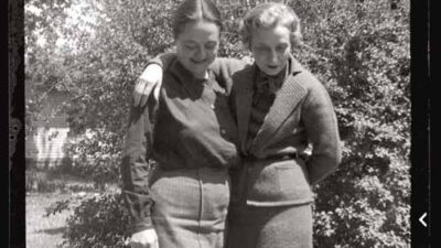 Two women stand outdoors with their arms around each other, both wearing long skirts and sweaters, in front of a bush and a house. [gen-ai]