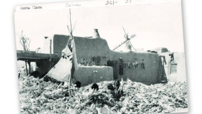Black-and-white photo of an adobe house with corn cobs spread out in the foreground and several people sitting among the corn; handwritten notes appear along the top edge of the photo. [gen-ai]