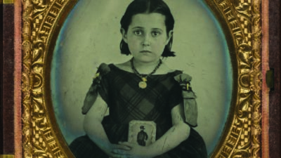 A young girl in a dark plaid dress sits for a formal portrait, holding a photo case, framed by an ornate gold oval frame and a decorative case. [gen-ai]