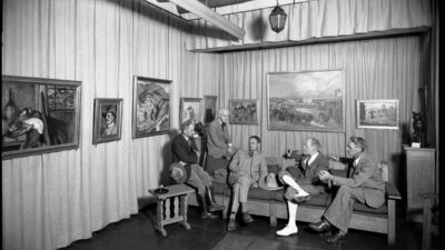 Five men in suits sit and stand in an art gallery, conversing. Several framed paintings and sculptures are displayed on the walls and furniture. [gen-ai]