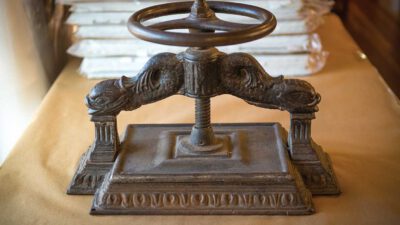 An antique cast iron book press with ornate detailing sits on a table, with stacks of wrapped items in the background. [gen-ai]