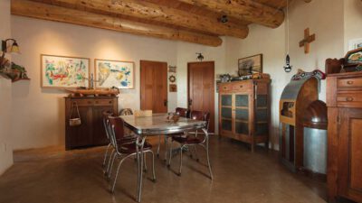 A rustic dining room with exposed wooden ceiling beams, a metal table with four chairs, wooden cabinets, art on the walls, and concrete flooring. [gen-ai]