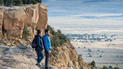 Two people with backpacks stand on a rocky cliff edge overlooking a vast valley and distant mountains under a partly cloudy sky. [gen-ai]