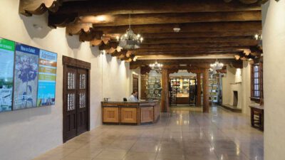 Spacious lobby with wooden ceiling beams, tiled floor, information desk, digital display screen, and a person seated at the counter. [gen-ai]