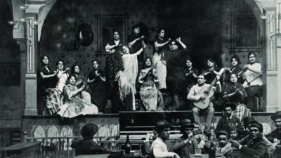Black-and-white photo of women performing onstage, some clapping, one with a guitar, while audience members sit at tables in the foreground, likely in a historic tavern or café setting. [gen-ai]